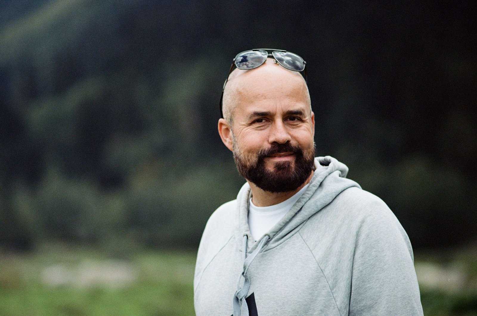 Portrait of a bald bearded man smiling outdoors wearing sunglasses and a hoodie. Warm and inviting.